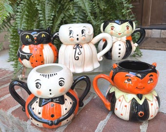 May include: A collection of five ceramic mugs shaped like Halloween characters, including a black cat, a white ghost, a vampire, a spider, and a devil. Each mug features a glossy finish, painted facial details, and a unique handle design.