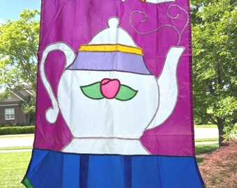 May include: A garden flag with a teapot and teacups design. The flag has a purple background with a white teapot, a pink rose, and three teacups with yellow accents. The bottom is blue with a green trim.