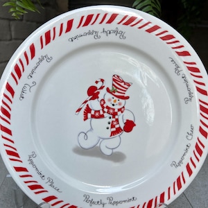 May include: White ceramic plate with a red and white candy cane stripe border. The center of the plate features a hand-painted snowman with a red hat, scarf, and mittens. The text "Perfectly Peppermint" and "Peppermint Place" are written on the plate.