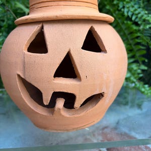 May include: A terracotta jack-o'-lantern with a matte finish, featuring triangular eyes, a triangular nose, and a wide, jagged mouth. The pumpkin is topped with a matching terracotta hat that has a flat crown and a wide, circular brim.
