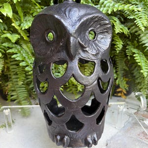 May include: A dark brown, cast metal owl-shaped lantern featuring a hollow, open-work body with circular cutouts. The owl has a textured surface, prominent eyes, and a small metal ring at the top for hanging. The design is symmetrical and rigid.