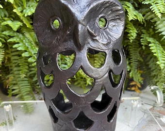 May include: A dark brown, cast metal owl-shaped lantern featuring a hollow, open-work body with circular cutouts. The owl has a textured surface, prominent eyes, and a small metal ring at the top for hanging. The design is symmetrical and rigid.