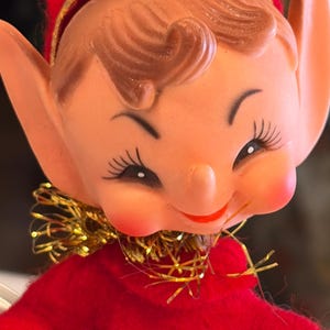 May include: A vintage elf figurine with a smiling face, rosy cheeks, and long eyelashes. The elf wears a red outfit with a gold collar and a red hat with a gold band. The elf's hair is brown.