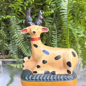 May include: A ceramic deer figurine with a tan body, black spots, and a red collar. The deer has black antlers and is resting on a green and yellow base. The background features lush green foliage.