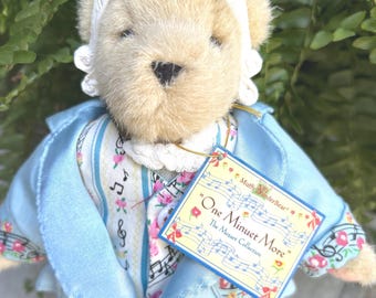 May include: A teddy bear dressed in a light blue jacket with floral and musical note patterns, a white bonnet, and light blue shoes. The bear has a tag that reads "One Minuet More" from The Mozart Collection.