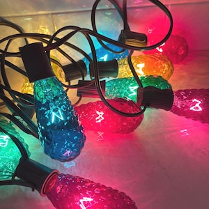 May include: A close-up of a string of colorful Christmas lights. The lights are various colors, including red, blue, green, and yellow. The bulbs are textured and have a vintage look. The black wire is visible.