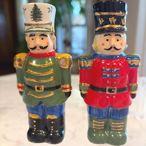 May include: Two ceramic nutcracker figurines. One is dressed in a green coat, blue pants, and a white hat with a Christmas tree. The other is in a red coat, blue pants, and a black hat. Both have mustaches and black boots.