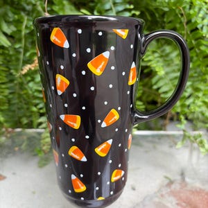 May include: A tall black ceramic mug featuring a repeating pattern of orange, yellow, and white candy corn shapes interspersed with small white polka dots. The mug has a smooth, glossy finish and a curved handle on the right side for holding.