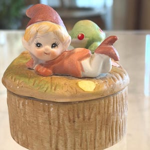 May include: A ceramic trinket box featuring a whimsical design. The box is topped with a figurine of a gnome in a red hat and orange outfit, with a green frog nearby. The box has a textured, wood-like appearance.