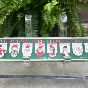 May include: Vintage Christmas card place settings box with illustrations of snowmen, Santa Claus, and angels. The box is green with red and white text, and the place settings are made in Japan.
