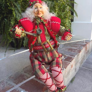 May include: A whimsical Christmas elf doll with a red hat and jacket, green shirt, and plaid pants. The doll has white curly hair, a smiling face, and is holding a pipe. The elf is wearing brown shoes and is decorated with holly.
