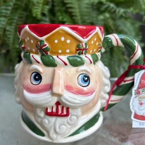 May include: A ceramic Christmas mug shaped like a nutcracker. The mug features a detailed face with blue eyes, a white beard, and a red mouth. The rim is decorated with a red and gold crown design, and the handle is a green and white candy cane. A tag is attached to the handle.