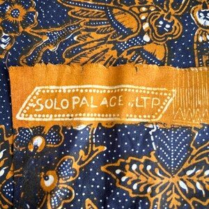 K&ouml;nnte beinhalten: Eine Nahaufnahme eines blauen und orangefarbenen Stoffes mit einem sich wiederholenden Muster aus Blumen und Bl&auml;ttern. Der Stoff hat ein Etikett mit der Aufschrift "SOLO PALACE ILTP".