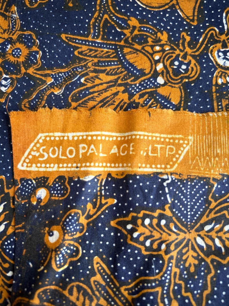 K&ouml;nnte beinhalten: Eine Nahaufnahme eines blauen und orangefarbenen Stoffes mit einem sich wiederholenden Muster aus Blumen und Bl&auml;ttern. Der Stoff hat ein Etikett mit der Aufschrift "SOLO PALACE ILTP".
