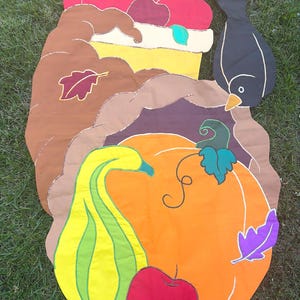 May include: A large, flat, fabric-based cornucopia decoration featuring a brown horn shape filled with a red apple, a yellow squash, a large orange pumpkin, and a black bird. The design includes colorful fabric leaves in red, green, and purple.