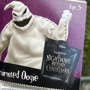 May include: A product tag for an animated Oogie Boogie toy from Tim Burton's The Nightmare Before Christmas. The tag has a purple background and features the character holding dice. The text "Age 3+" is visible.