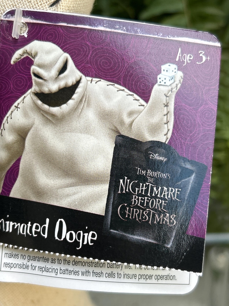 May include: A product tag for an animated Oogie Boogie toy from Tim Burton's The Nightmare Before Christmas. The tag has a purple background and features the character holding dice. The text "Age 3+" is visible.
