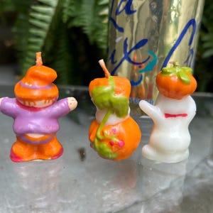 K&ouml;nnte beinhalten: Drei Halloween-Kerzen. Eine ist ein lila und orangefarbener Zwerg, eine weitere ein orangefarbener K&uuml;rbis mit gr&uuml;nen Akzenten und die dritte ein wei&szlig;es Gespenst mit roter Schleife. Die Kerzen stehen auf einer Glasoberfl&auml;che.