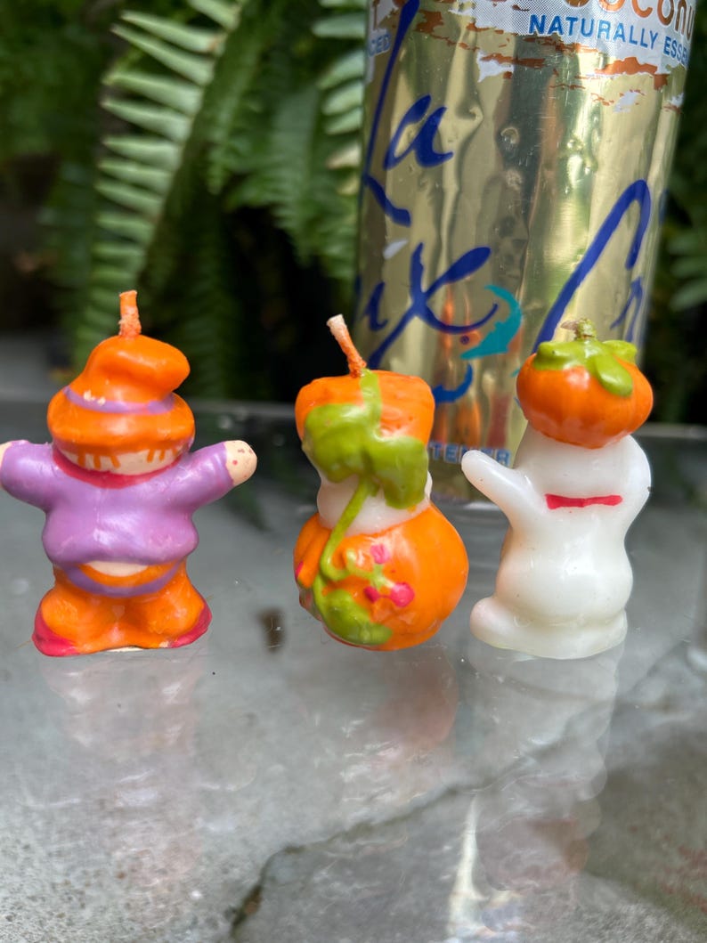 K&ouml;nnte beinhalten: Drei Halloween-Kerzen. Eine ist ein lila und orangefarbener Zwerg, eine weitere ein orangefarbener K&uuml;rbis mit gr&uuml;nen Akzenten und die dritte ein wei&szlig;es Gespenst mit roter Schleife. Die Kerzen stehen auf einer Glasoberfl&auml;che.