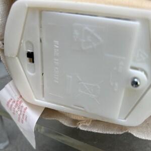 May include: A close-up of a white plastic battery compartment with a black on/off switch. The compartment is labelled "MADE IN CHINA" and features recycling symbols. A fabric-covered object is visible.