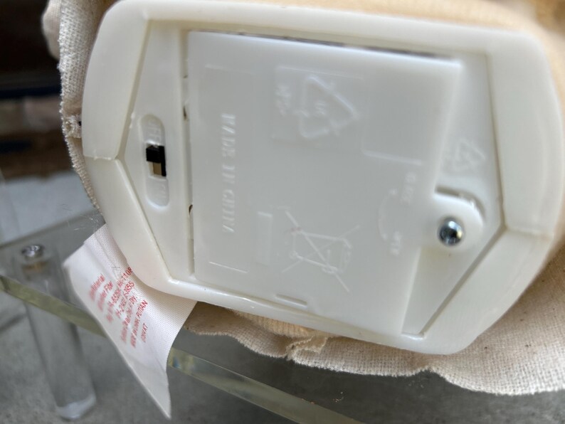 May include: A close-up of a white plastic battery compartment with a black on/off switch. The compartment is labelled "MADE IN CHINA" and features recycling symbols. A fabric-covered object is visible.