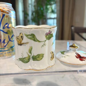 May include: A white porcelain jar with a lid, featuring hand-painted butterflies, green leaves, and pink flowers. The jar has a gold trim and a scalloped edge. The lid is off to the side.