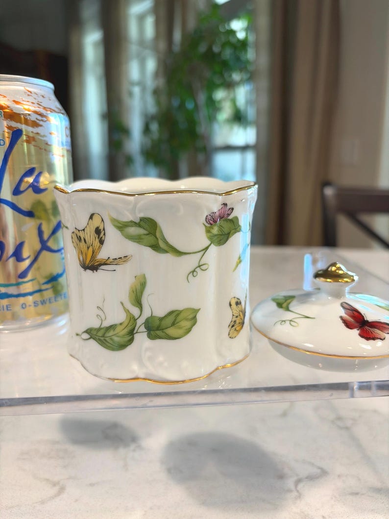 May include: A white porcelain jar with a lid, featuring hand-painted butterflies, green leaves, and pink flowers. The jar has a gold trim and a scalloped edge. The lid is off to the side.