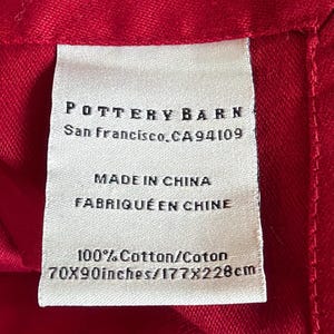 May include: A red textile with a white label. The label reads "POTTERY BARN, San Francisco, CA 94109, MADE IN CHINA, 100% Cotton, 70x90 inches". The fabric is a solid, vibrant red color.