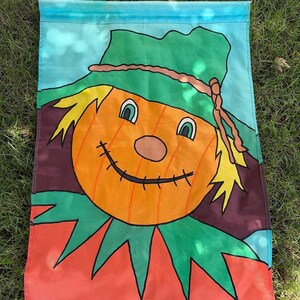 May include: A rectangular garden flag featuring a cartoon scarecrow with a round orange pumpkin head, a green hat with a brown rope band, yellow hair, and a stitched smile. The background includes a blue sky and a red base with green leaf accents.
