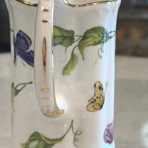 May include: White porcelain pitcher with a gold rim and handle. The pitcher is decorated with colorful butterflies and green leaves. The handle has gold accents. A delicate and elegant design.
