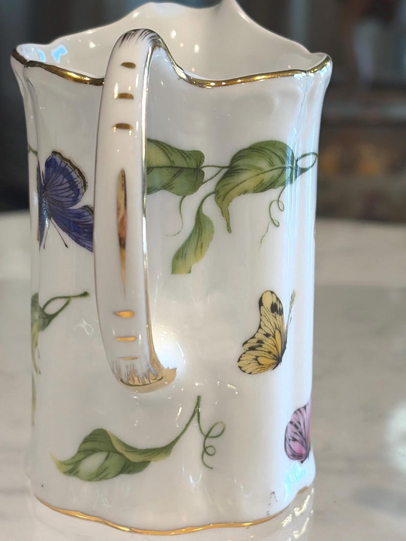 May include: White porcelain pitcher with a gold rim and handle. The pitcher is decorated with colorful butterflies and green leaves. The handle has gold accents. A delicate and elegant design.