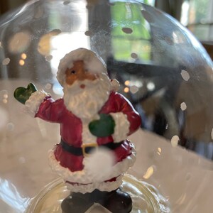 May include: A glass snow globe with a Santa Claus figurine inside. The globe is filled with white glitter and has a white base. The Santa Claus figurine is wearing a red suit with white trim and a white beard. He is holding a green bag in his left hand and a white ball in his right hand.