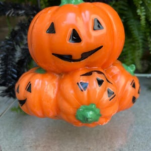 May include: A stack of orange ceramic jack-o'-lanterns with black cut-out eyes, noses, and mouths. The top pumpkin has a green stem. The bottom pumpkins have green stems and smiling faces. A festive Halloween decoration.