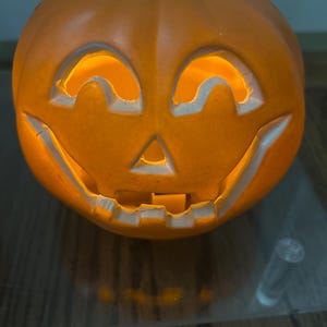 May include: Orange plastic jack-o'-lantern with a carved face and a light inside. The pumpkin has a black stem and is sitting on a reflective surface. A classic Halloween decoration.