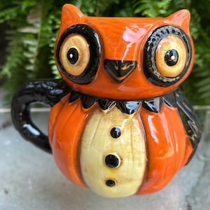 May include: Orange owl-shaped ceramic mug with black accents. The mug features large, round eyes, a black beak, and a yellow and black bow tie design. The owl's chest is a light yellow color with three black buttons. The handle and wings are black.