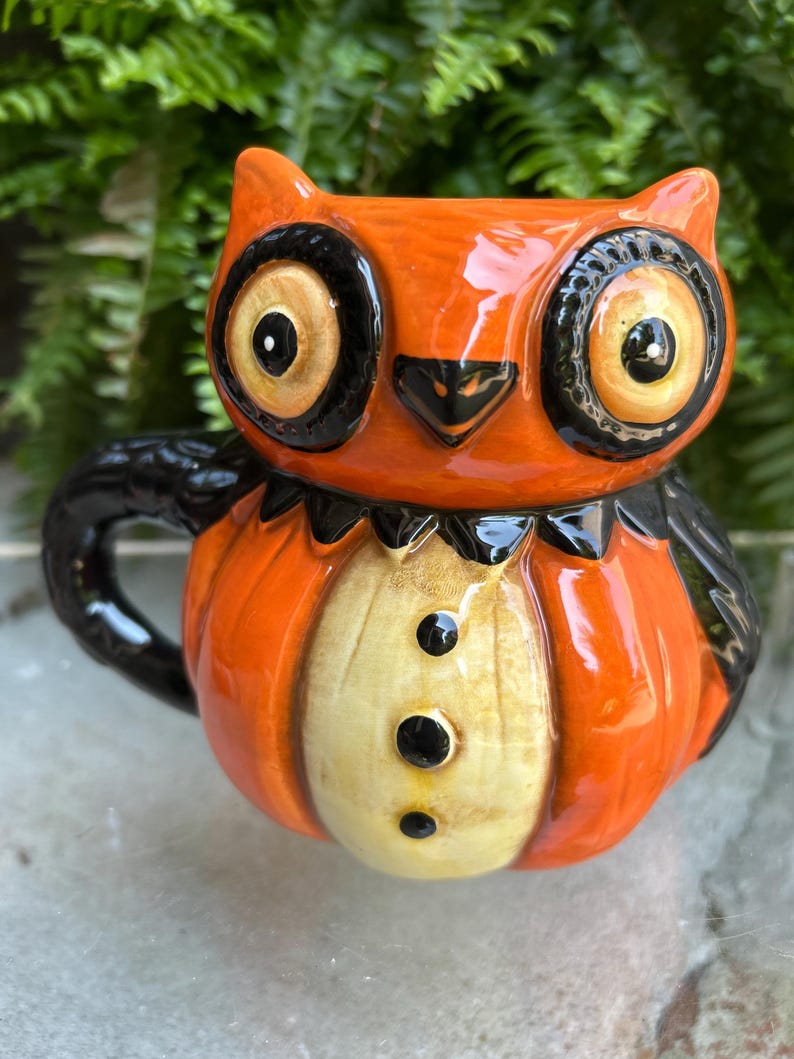 May include: Orange owl-shaped ceramic mug with black accents. The mug features large, round eyes, a black beak, and a yellow and black bow tie design. The owl's chest is a light yellow color with three black buttons. The handle and wings are black.