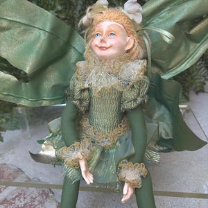 May include: A decorative fairy doll with curly blonde hair, blue eyes, and a pointed nose. The doll wears a green textured dress with gold-trimmed ruffled collar and cuffs, featuring large, translucent green wings attached to the back.