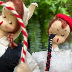 May include: Two vintage doll figures with closed eyes. One doll wears a red beret and holds a black and red striped object. The other doll has braided hair and a black and white outfit, holding a red and white striped rope.