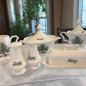 May include: A white ceramic Christmas-themed serving set. The set includes a creamer, sugar bowl, salt and pepper shakers, a covered serving dish, and a butter dish. Each piece features a Christmas tree design and holly accents.