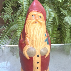 May include: A tall, conical figurine of a bearded figure wearing a deep red pointed hat and matching coat with a tan trim. The figure has a long, textured tan beard, black buttons down the front, and holds a small tan animal and a blue bird in its hands.