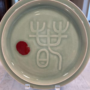 May include: A light green ceramic plate with a raised design of a stylized tree and a red circle. The plate has a white rim and is slightly raised in the center.