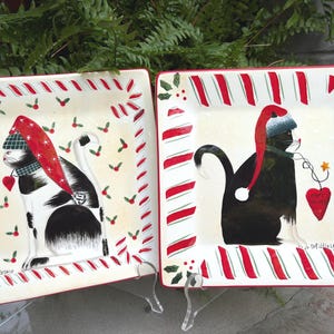 May include: Two square ceramic plates featuring black and white cats wearing red Santa hats. The borders are decorated with red and white candy cane stripes and green holly leaves. One cat wears a plaid scarf, while the other holds a red heart ornament.