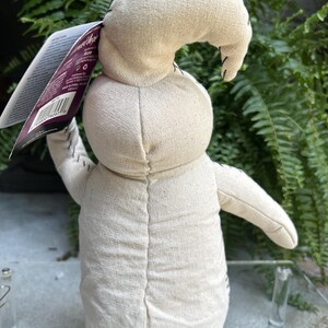 May include: A beige fabric doll with a stitched design, featuring a curved head and outstretched arms. The doll has black stitching details and a tag attached. The background includes green foliage.