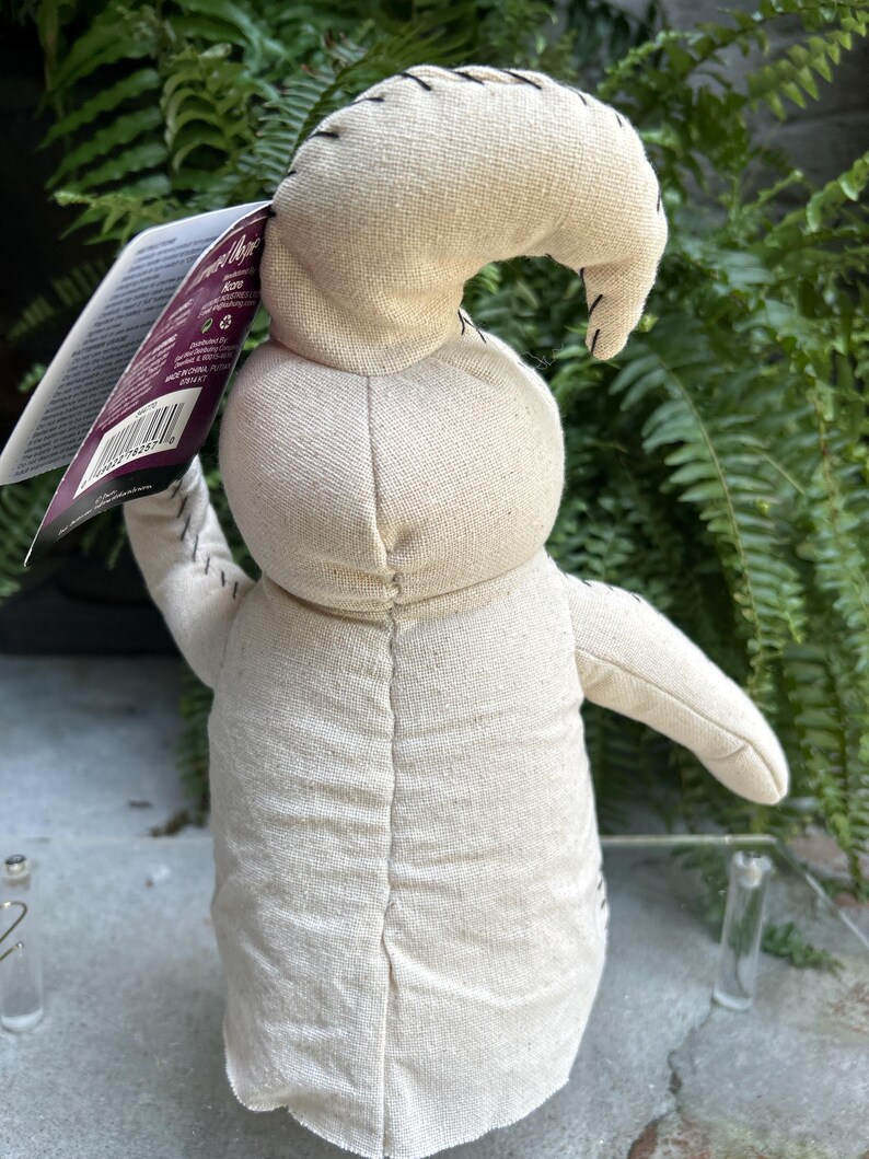 May include: A beige fabric doll with a stitched design, featuring a curved head and outstretched arms. The doll has black stitching details and a tag attached. The background includes green foliage.