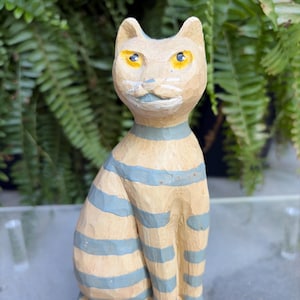 May include: A carved wooden cat figurine with a light tan body and blue horizontal stripes. The cat has yellow eyes, a blue collar, and a painted mustache. The cat is sitting upright with its legs together.