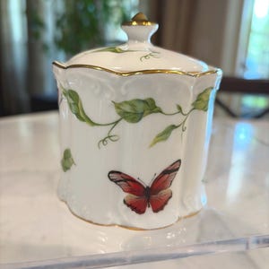 May include: A white porcelain jar with a lid, adorned with green leaf vines and a red butterfly. The jar has gold trim and a gold knob on the lid. A decorative piece for the home.