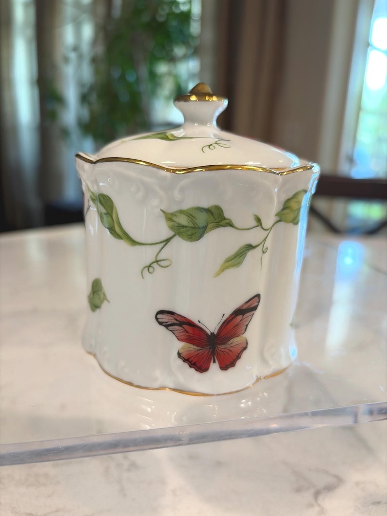 May include: A white porcelain jar with a lid, adorned with green leaf vines and a red butterfly. The jar has gold trim and a gold knob on the lid. A decorative piece for the home.