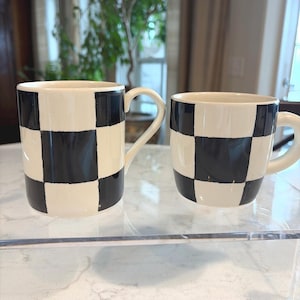 May include: Two ceramic mugs with a black and white checkered pattern. The mugs have a cream-colored base and a curved handle. The mugs are displayed on a clear surface, with a blurred background of a room with a plant.