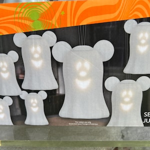 May include: Set of 8 Halloween ghost decorations. Each white ghost has a smiling jack-o'-lantern face and mouse-ear-shaped head. The ghosts are lit from within, creating a spooky glow. The packaging has an orange and black Halloween theme.