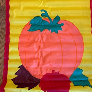 May include: A decorative fall-themed flag featuring a large orange pumpkin with green leaves and a red stem. The pumpkin is set against a yellow background with red borders, creating a festive autumn display.